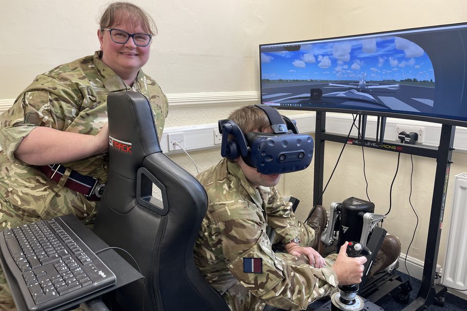 Woman adult volunteer oversees another volunteer at the controls of the virtual reality aircraft simulator