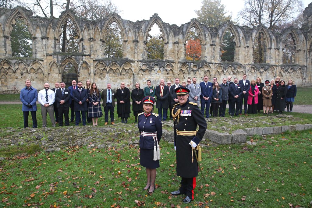 Man and woman in military uniform in front of a group of people in civilian clothing against a backdrop of the ruins of an ancient building