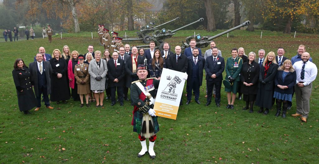 A large group of people wrapped in warm coats in front of soldiers and large artillery guns with one soldier in kilt and scottish military hat and sporran carrying a Gold Award sign