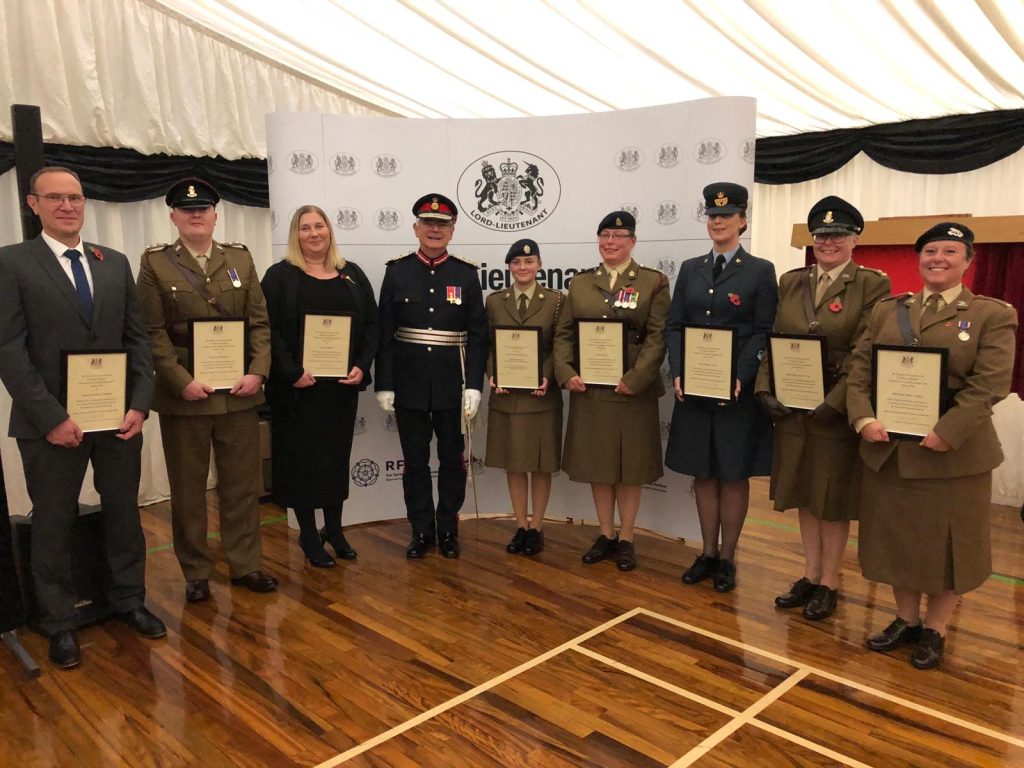 Line of nine people, all but two wearing military uniform and holding certificates. In the centre is Lord-Lieutenant - a man in quasi military uniform smiling at the camera