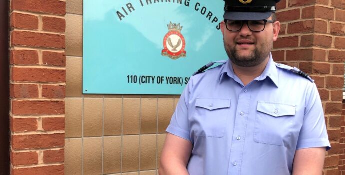 Gianluca Gaudiso standing in his air cadet volunteer uniform in front of a sign which says Air Training Corps, 110 City of York Squadron