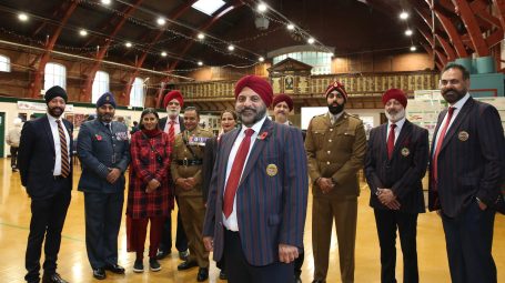 Man in turban smiles with members of the Armed Forces also dressed in turbans and members of the community in high-ceilinged building