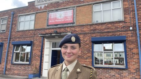 Female army cadet in dress uniform smiling, standing in front of red-brick Middleton Barracks.