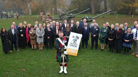 A large group of people wrapped in warm coats in front of soldiers and large artillery guns with one soldier in kilt and scottish military hat and sporran carrying a Gold Award sign